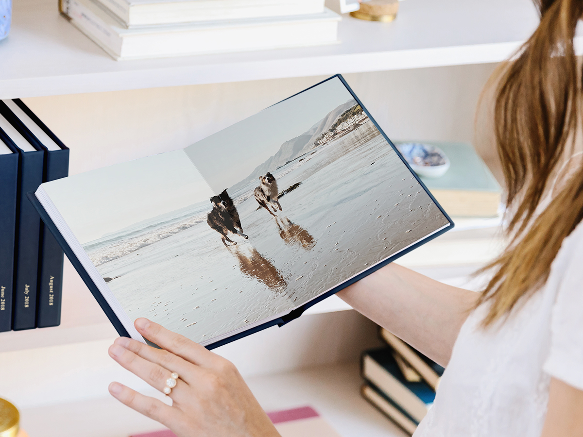 Woman holding up Everyday Photo Book opened up to a two-page spread of Australian Shepherds running along the beach