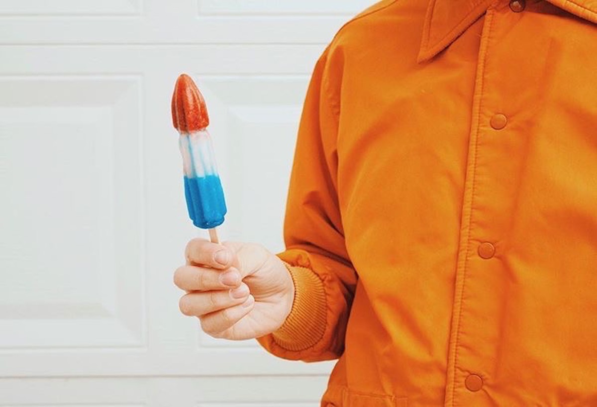 A red, white and blue ice popsicle held by a person in an orange button-up shirt