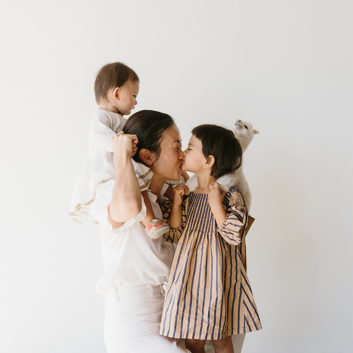 Mom with baby on her shoulder kissing toddler while making a funny face