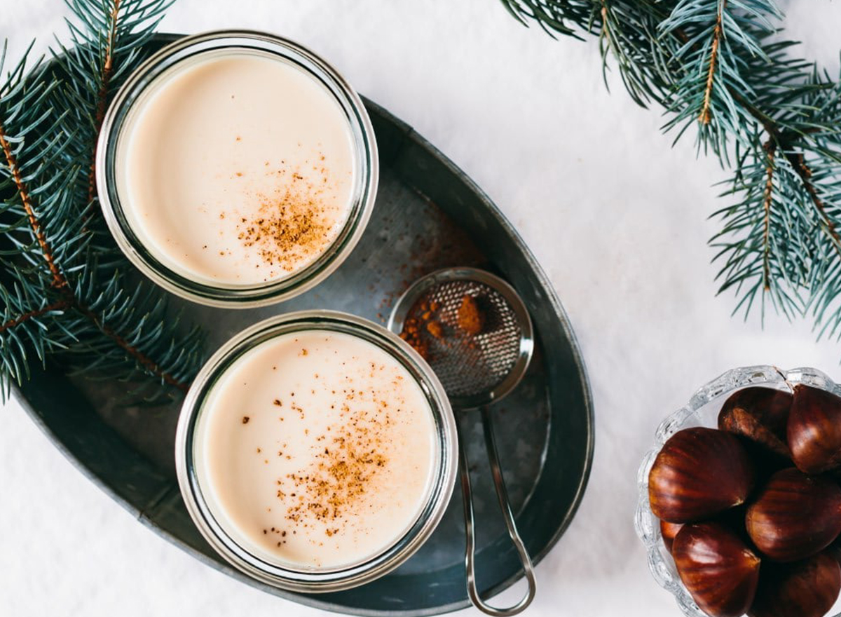 two cups of eggnog on serving tray next to sprig of pine and bowl of chestnuts